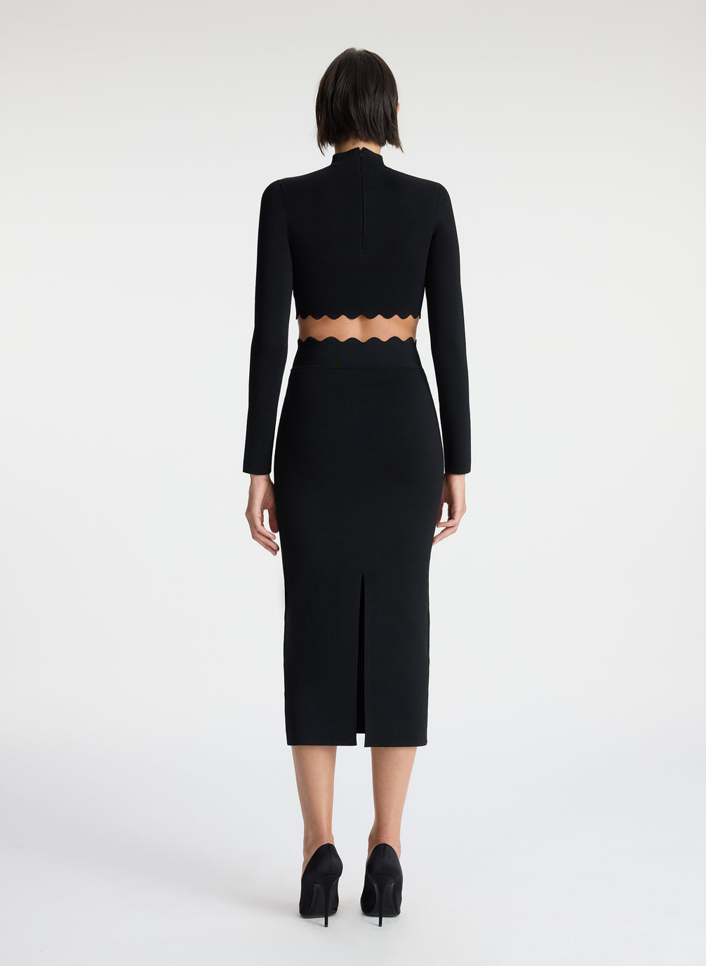 back view of woman wearing black long sleeve crop top with scallop detailing and matching black scalloped detailing knit midi skirt