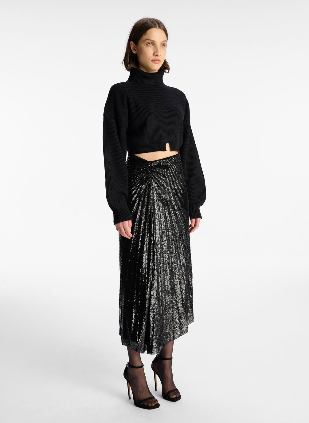 side  view of a woman wearing a black cropped turtleneck sweater and black sequined pleated midi skirt