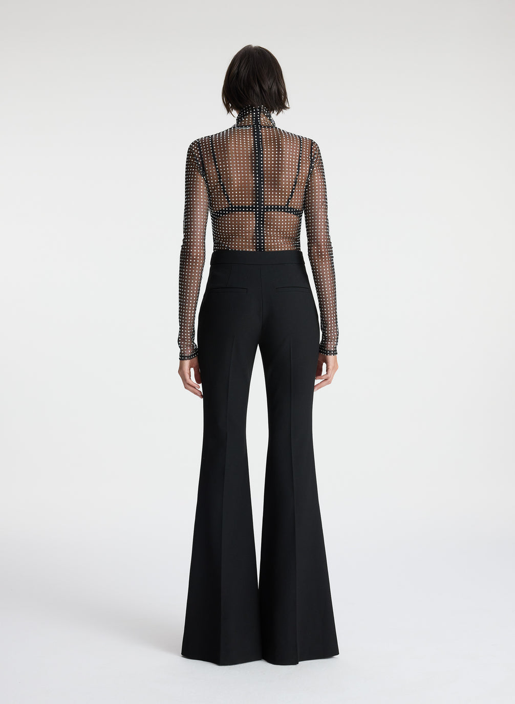 back view of woman wearing sheer black mock neck long sleeve with allover rhinestone embellishment and black pants