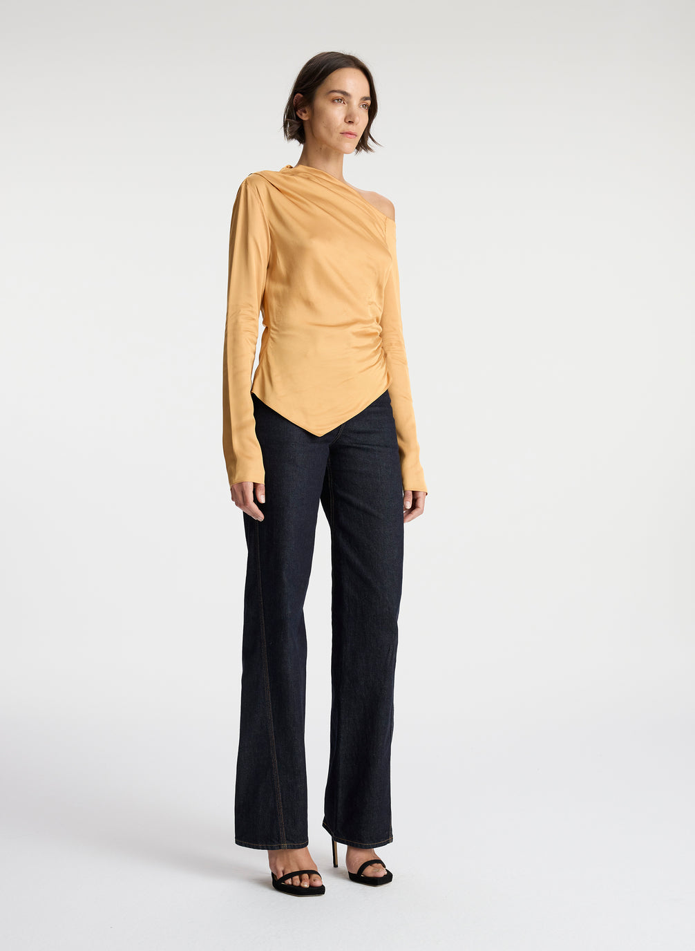 side view of woman wearing asymmetric gold satin long sleeve top and dark wash denim jeans