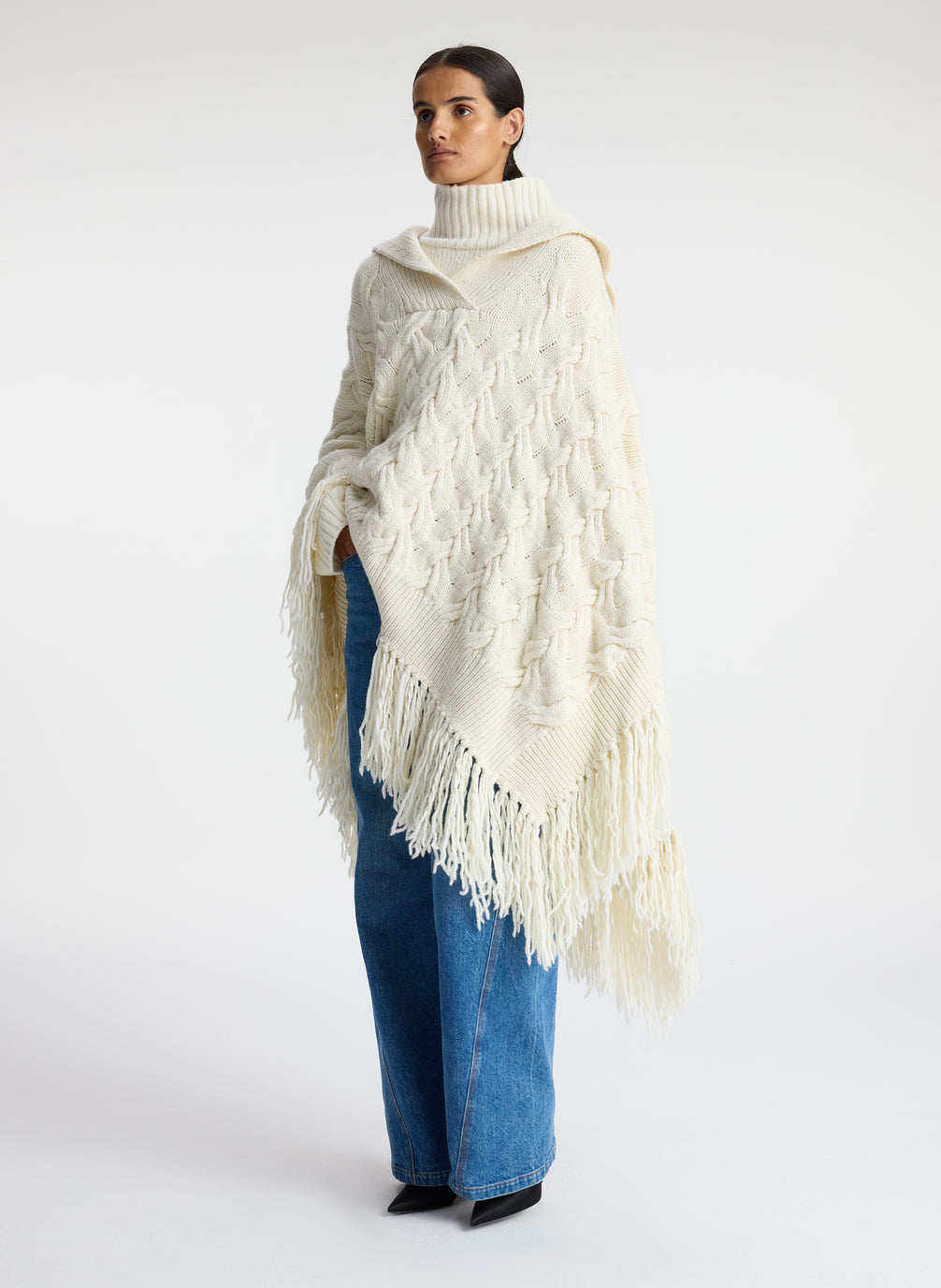 side view of woman wearing cream cable knit poncho with fringe