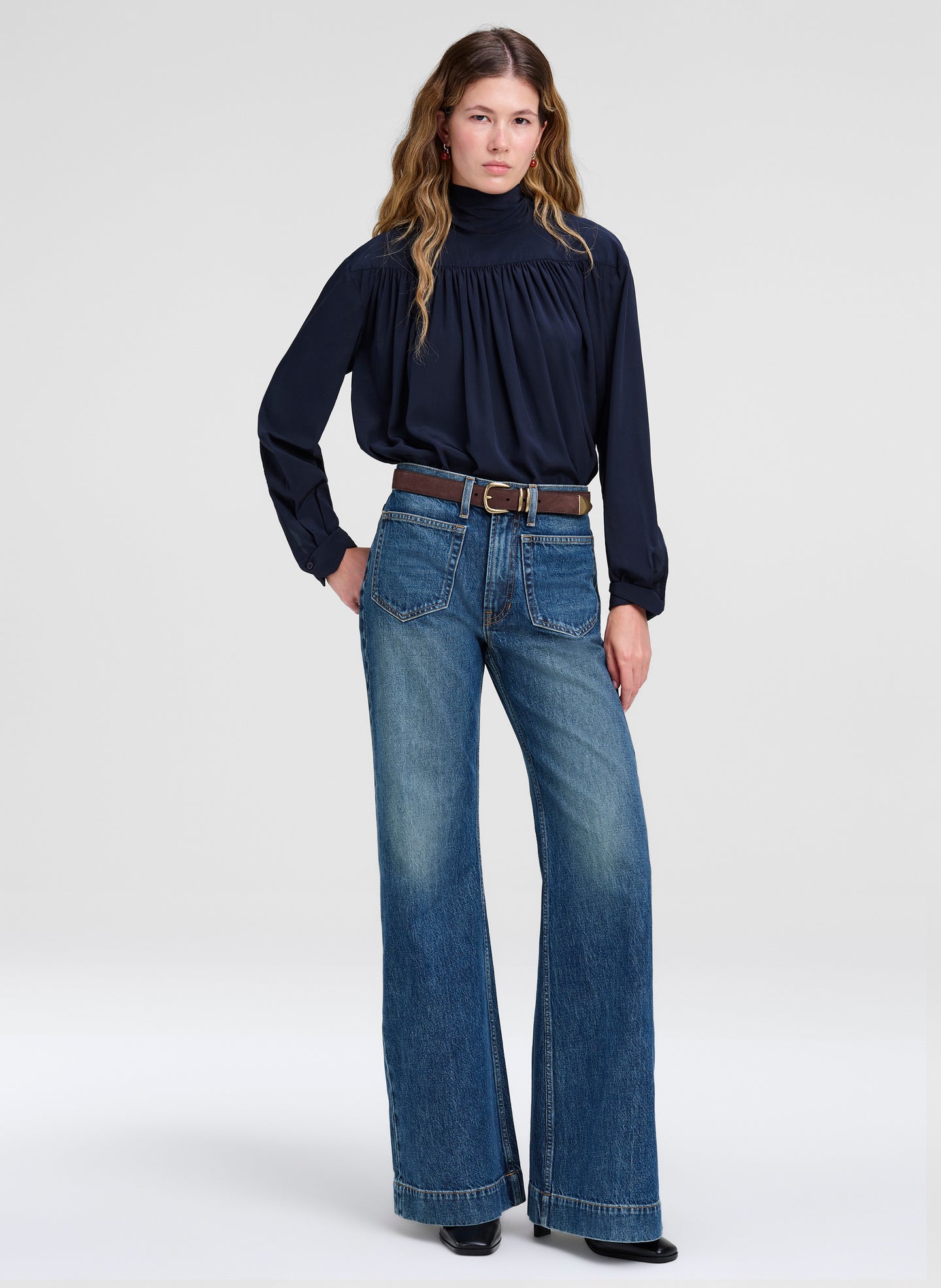 A model wearing a dark navy blue long-sleeve blouse with a gathered turtleneck and wide-leg blue jeans, styled with a brown belt, standing against a light gray studio background.