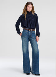 A model wearing a dark navy blue long-sleeve blouse with a gathered turtleneck and wide-leg blue jeans, styled with a brown belt, standing against a light gray studio background.