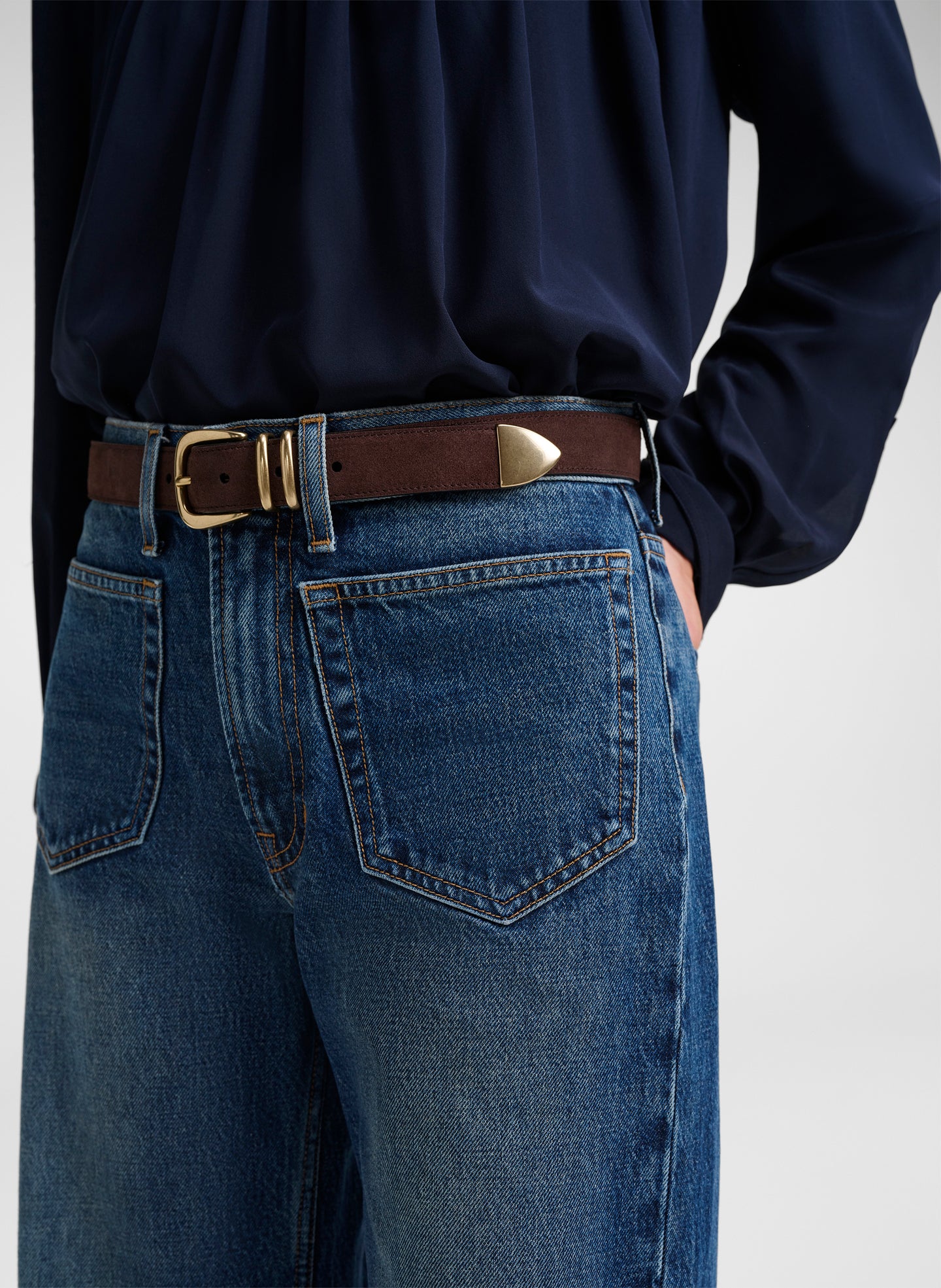Close-up of a person wearing blue jeans with a brown leather belt featuring gold-tone hardware, paired with a dark navy top against a light gray background.