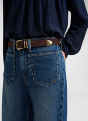 Close-up of a person wearing blue jeans with a brown leather belt featuring gold-tone hardware, paired with a dark navy top against a light gray background.