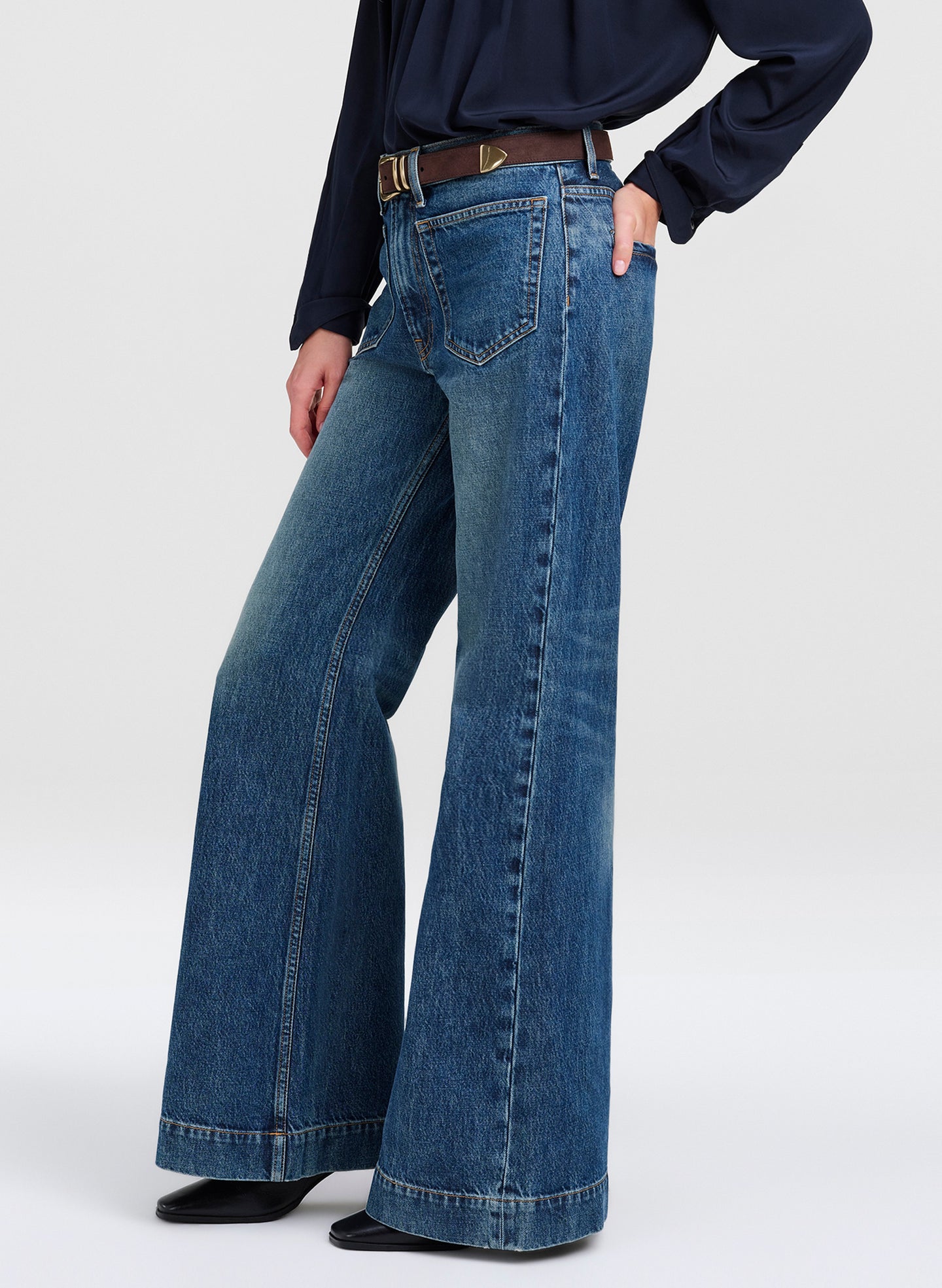A person wearing blue wide-leg denim jeans with a dark navy long-sleeve top and a brown belt, shown from the waist down against a light gray studio background.