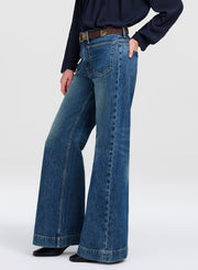 A person wearing blue wide-leg denim jeans with a dark navy long-sleeve top and a brown belt, shown from the waist down against a light gray studio background.