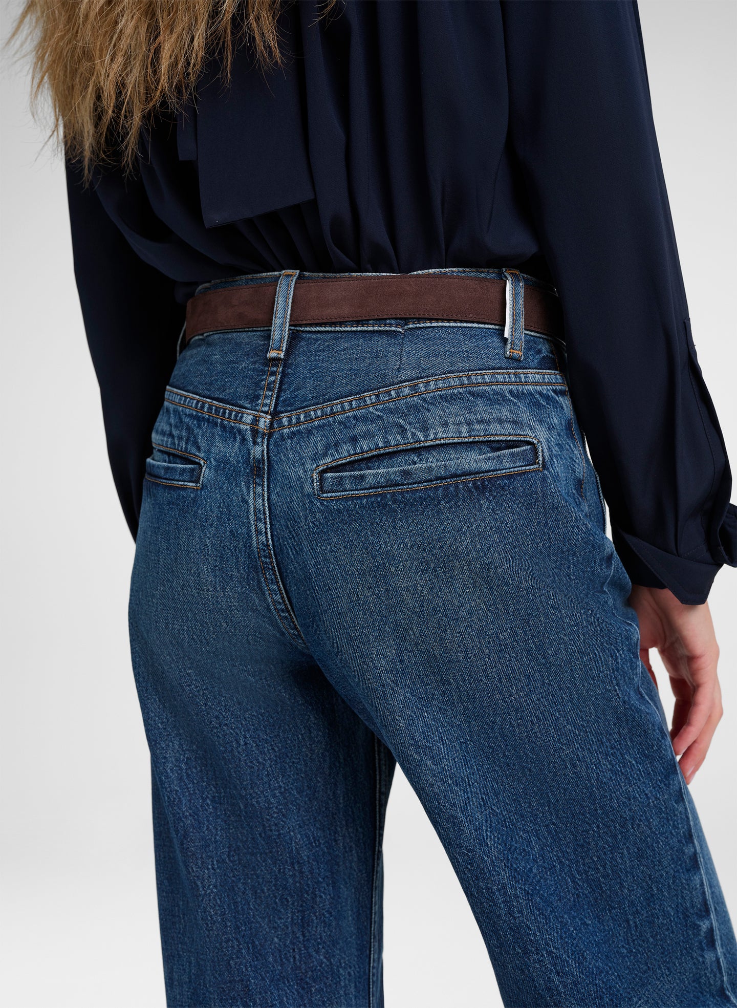 Close-up back view of a person wearing blue denim jeans with a brown belt and a dark navy blouse, against a light gray studio background.
