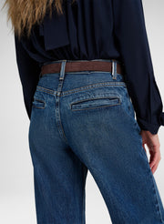 Close-up back view of a person wearing blue denim jeans with a brown belt and a dark navy blouse, against a light gray studio background.