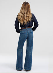 A woman viewed from behind wearing blue wide-leg flare jeans, a dark navy long-sleeve top, and a brown belt, standing against a light gray studio background.