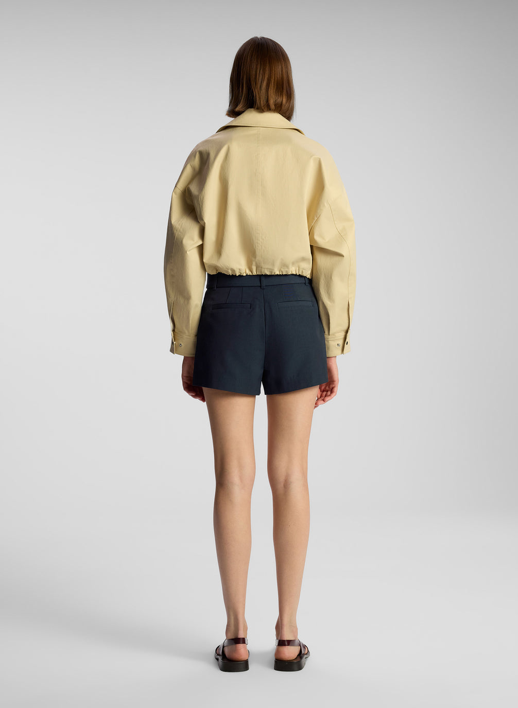 back view of woman wearing tan jacket, cream bodysuit, and navy blue shorts