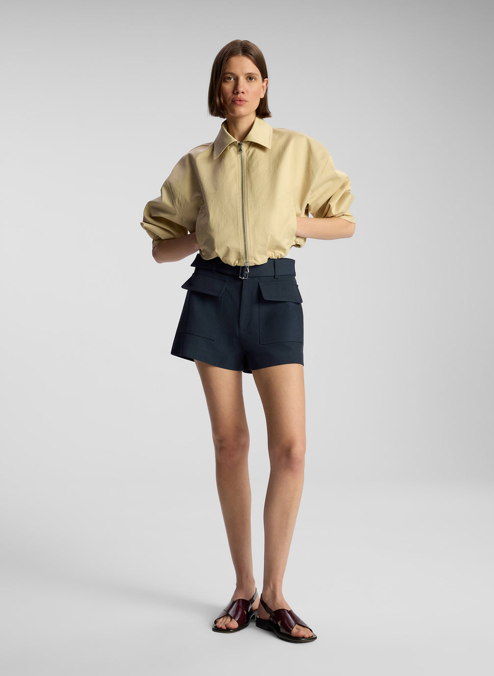 front view of woman wearing tan jacket, cream bodysuit, and navy blue shorts