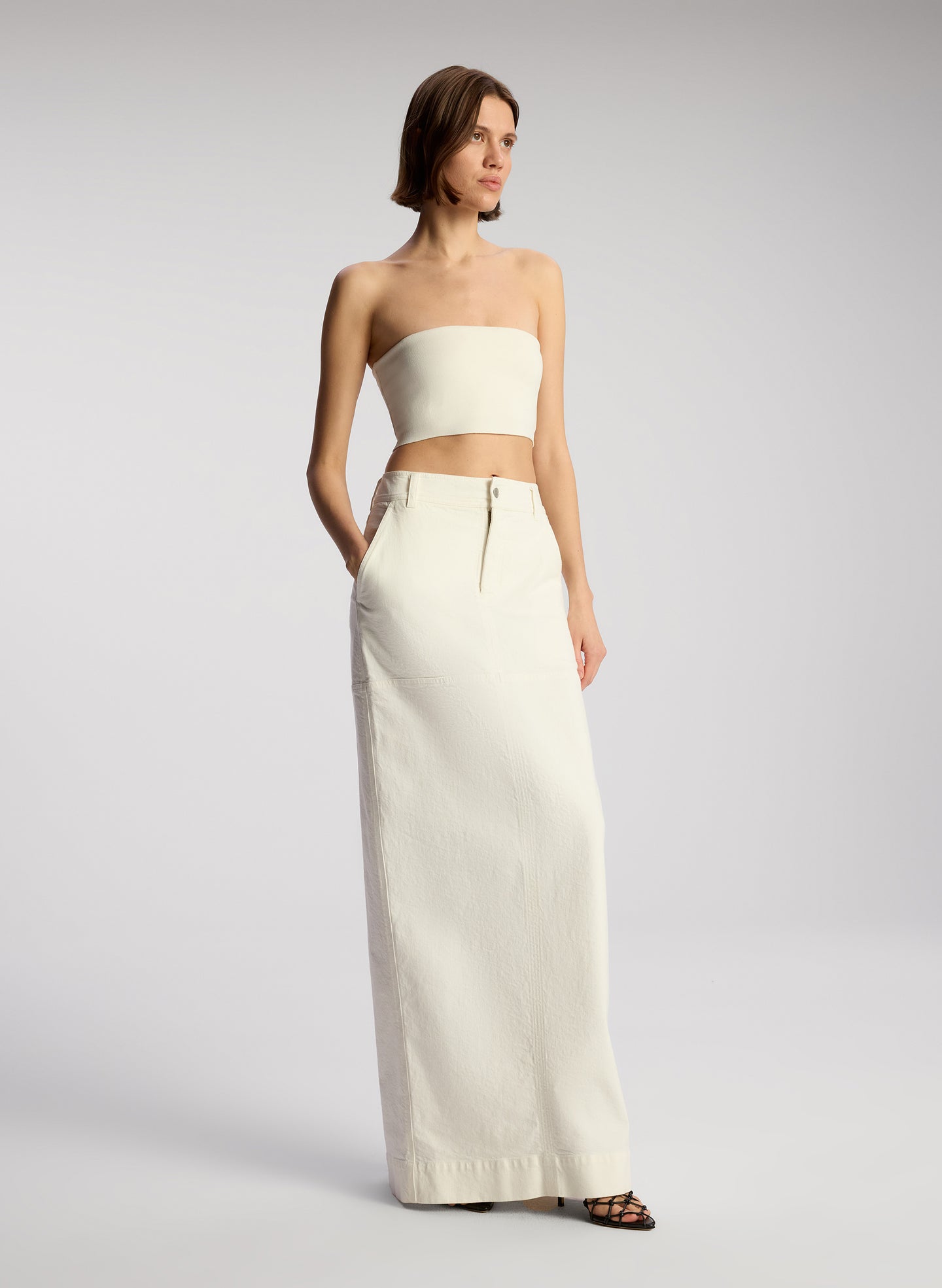 side view of woman wearing off white cropped strapless top with white maxi skirt