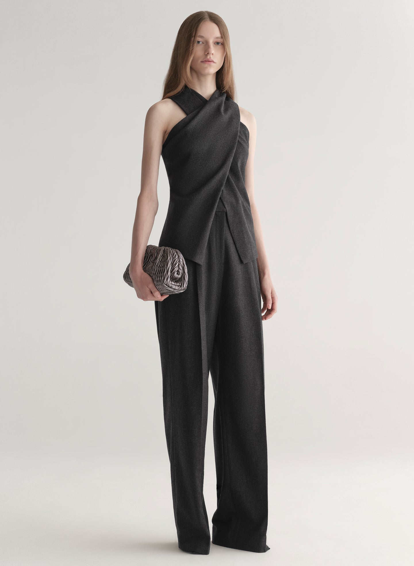 Woman wearing a grey draped top and pants holding a clutch on a white background