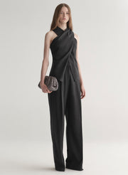 Woman wearing a grey draped top and pants holding a clutch on a white background