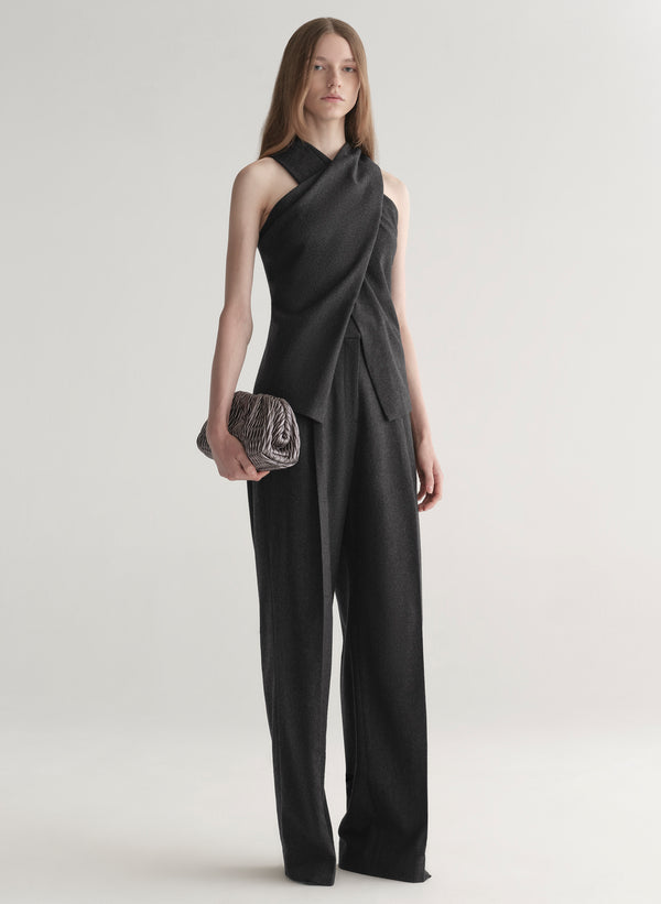 Woman wearing a grey draped top and pants holding a clutch on a white background