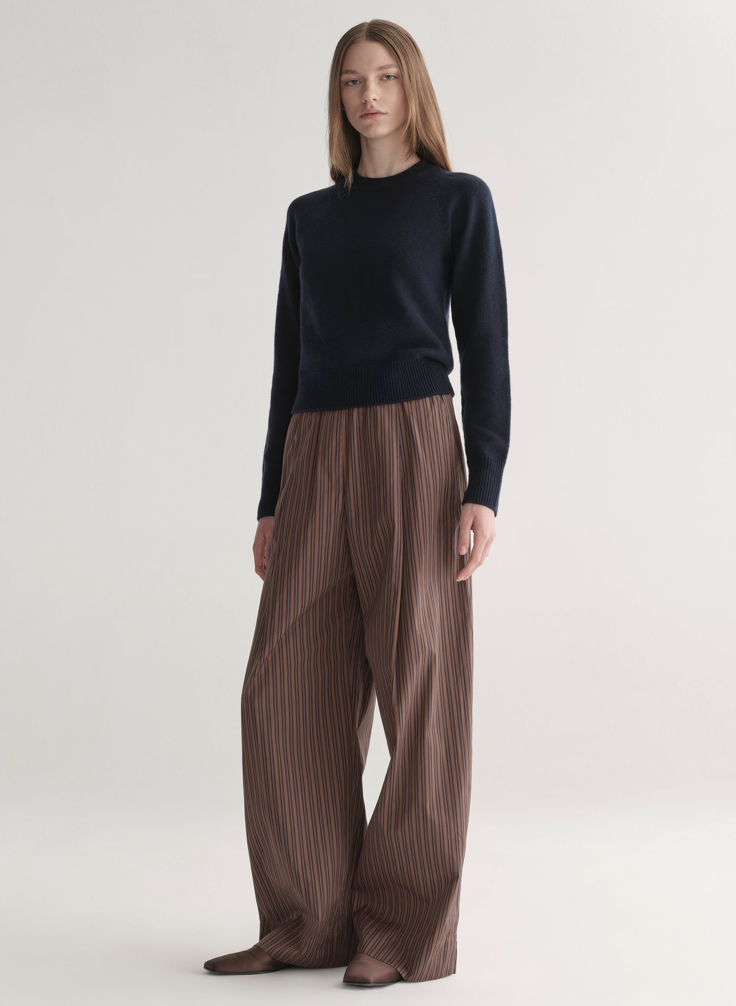 Person wearing a navy blue sweater and brown pleated pants on a white background
