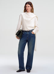 A model stands against a light gray studio background wearing a cream draped long-sleeve blouse, blue straight-leg jeans, black shoes, and holding a small black handbag.