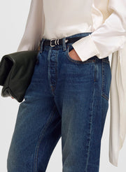 A close-up of a person wearing blue denim jeans with a black belt, a white blouse, and holding a black clutch bag against a light gray background.