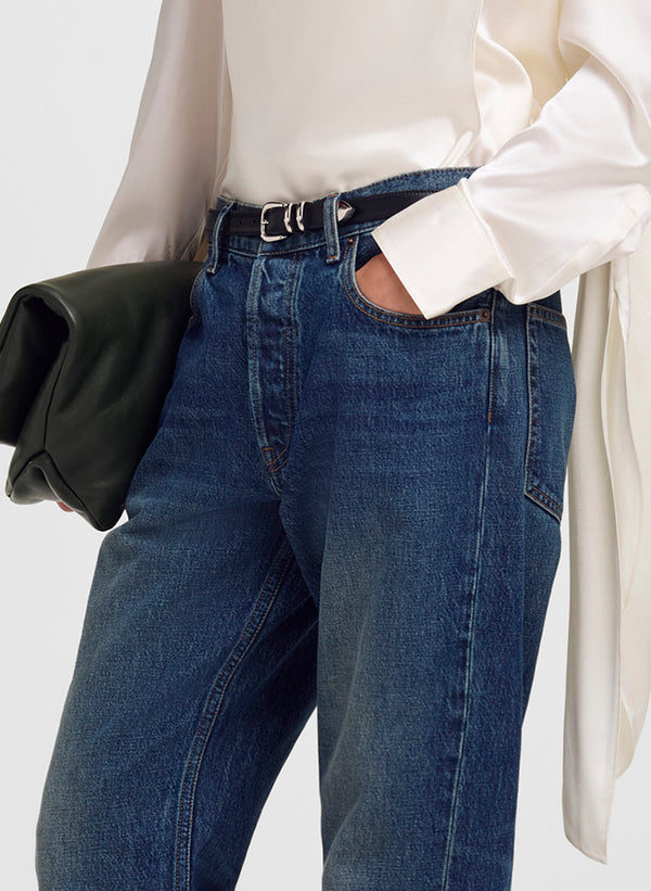 A close-up of a person wearing blue denim jeans with a black belt, a white blouse, and holding a black clutch bag against a light gray background.