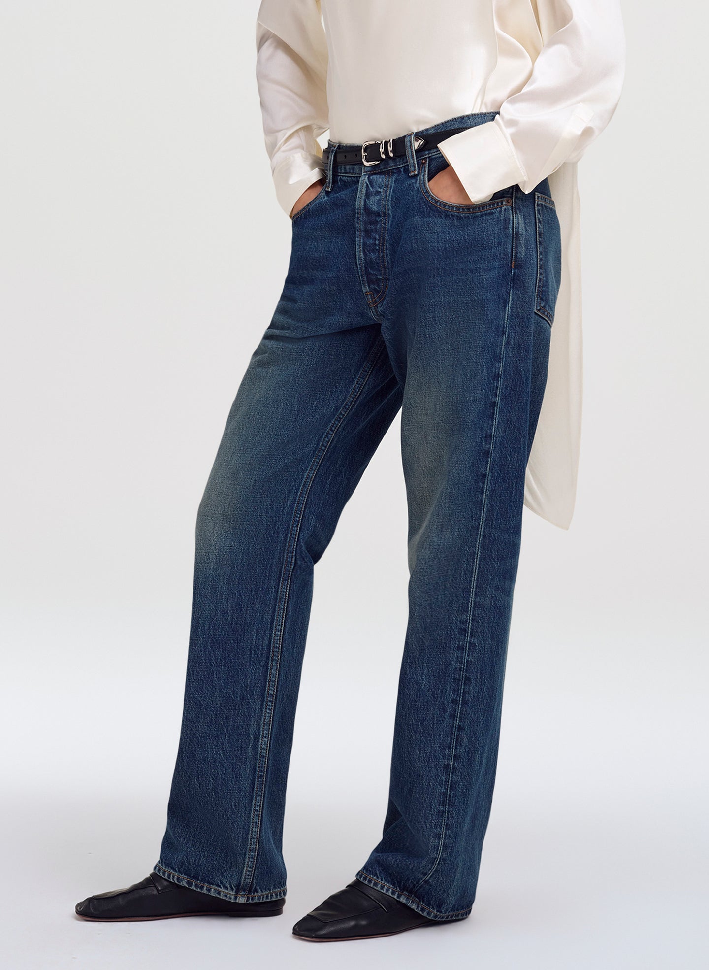 A person wearing dark blue straight-leg denim jeans with a black belt, paired with a cream satin shirt and black shoes, shown from the waist down against a light gray studio background.