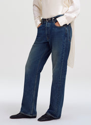 A person wearing dark blue straight-leg denim jeans with a black belt, paired with a cream satin shirt and black shoes, shown from the waist down against a light gray studio background.