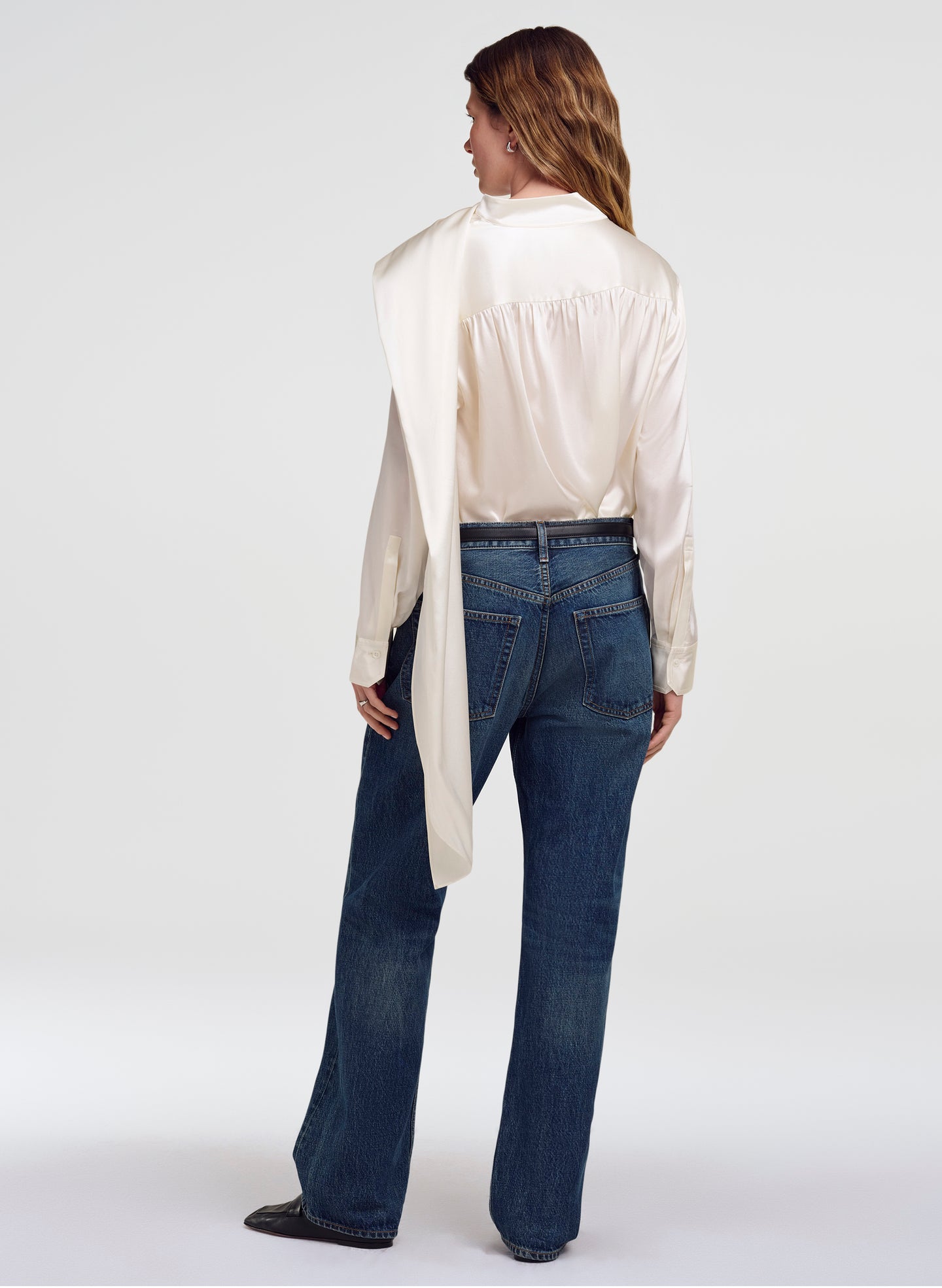 A woman shown from the back wearing a cream satin shirt with a draped collar and dark blue jeans, photographed against a light gray studio background.