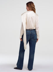 A woman shown from the back wearing a cream satin shirt with a draped collar and dark blue jeans, photographed against a light gray studio background.