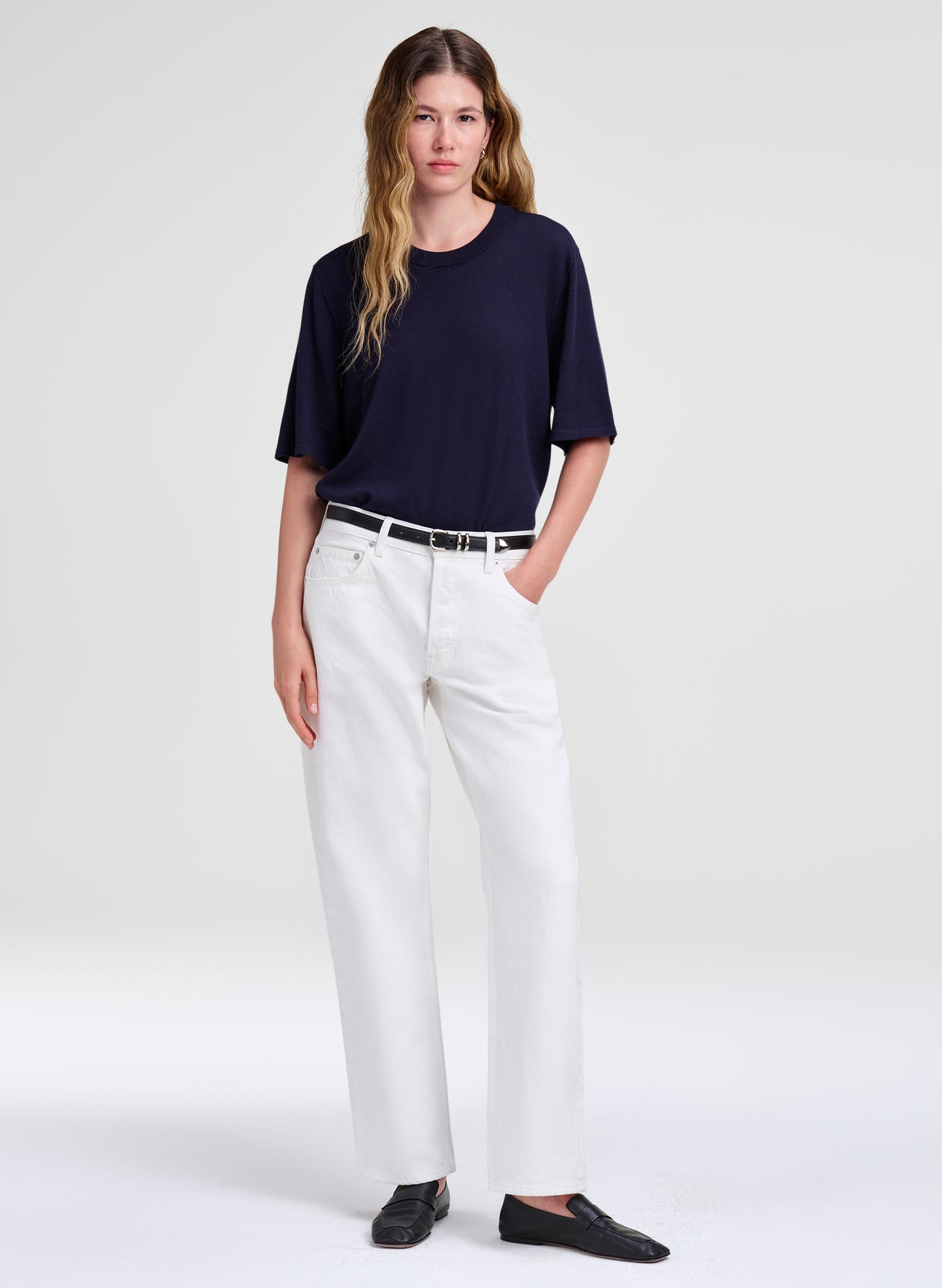 A woman modeling a dark navy short-sleeve crewneck top, white wide-leg trousers, a thin black belt, and black slip-on shoes against a light gray studio background.