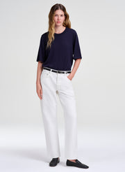 A woman modeling a dark navy short-sleeve crewneck top, white wide-leg trousers, a thin black belt, and black slip-on shoes against a light gray studio background.