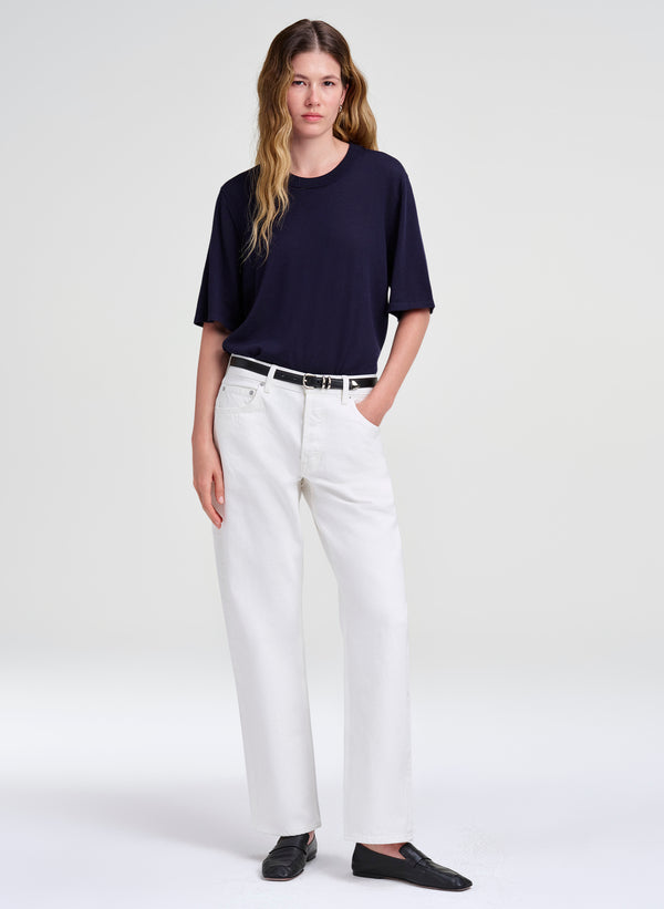 A woman modeling a dark navy short-sleeve crewneck top, white wide-leg trousers, a thin black belt, and black slip-on shoes against a light gray studio background.