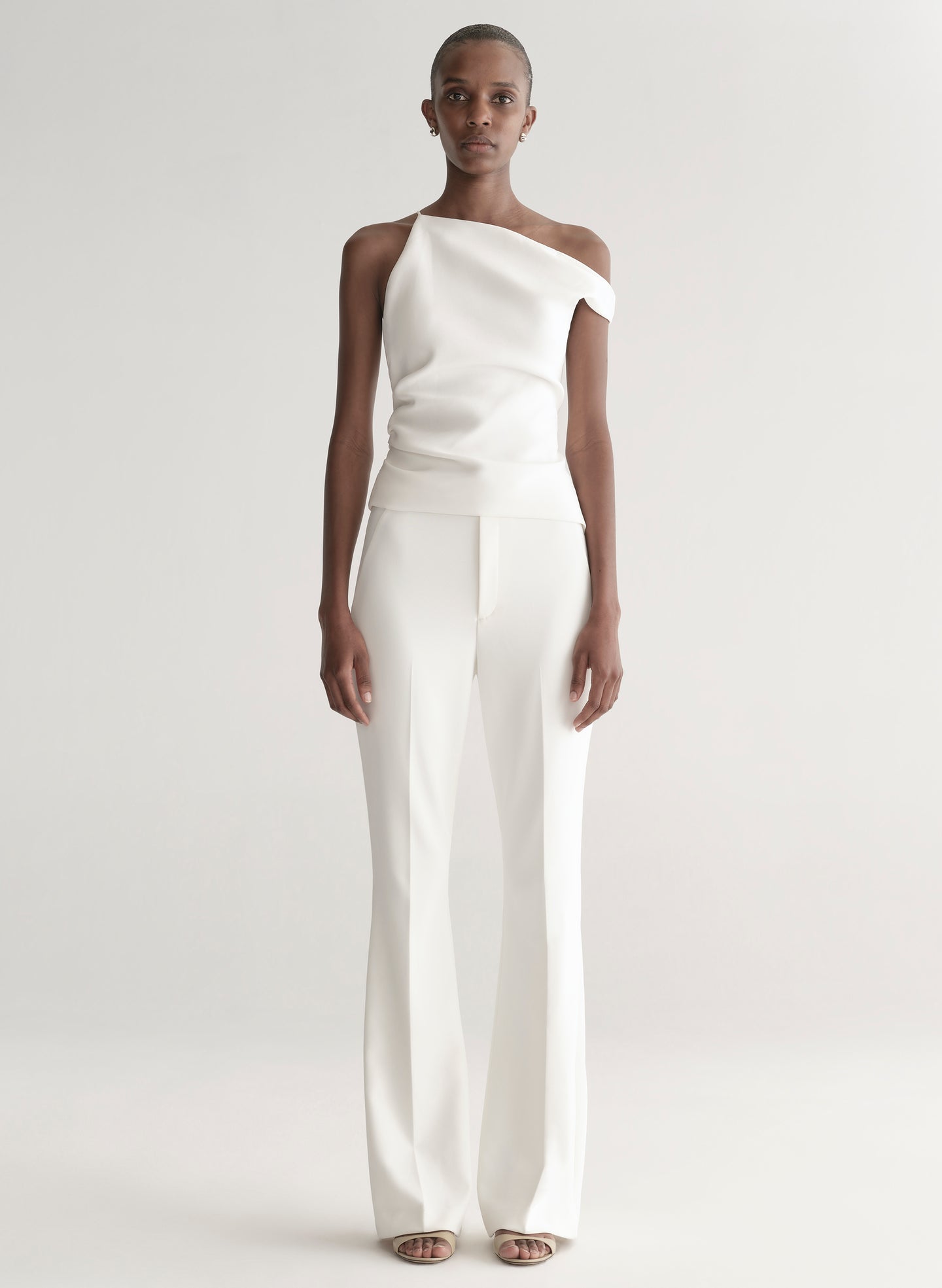 Woman wearing a white one-shoulder top and white pants on a plain background