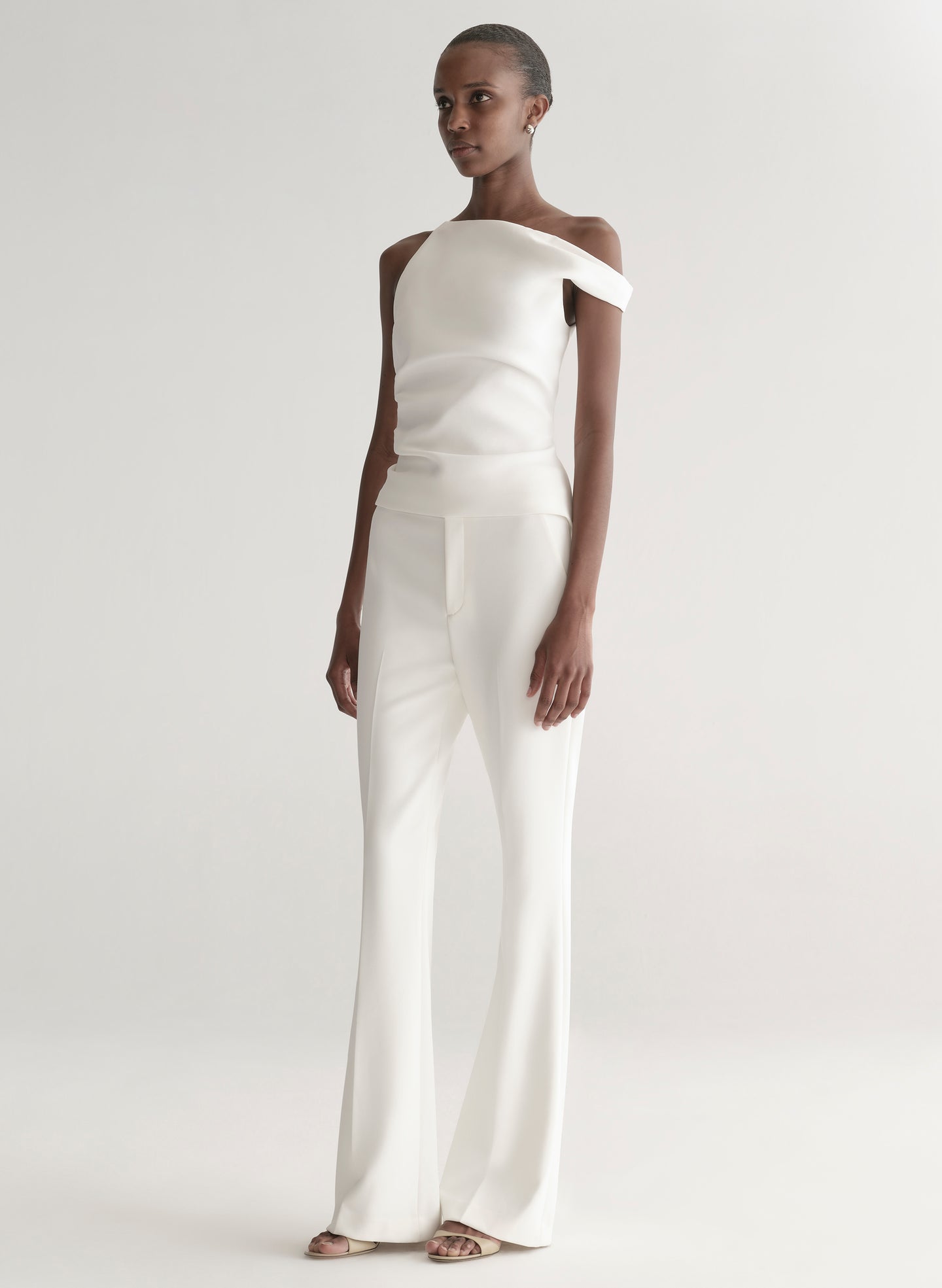 Woman wearing a white one-shoulder top and white pants on a plain background
