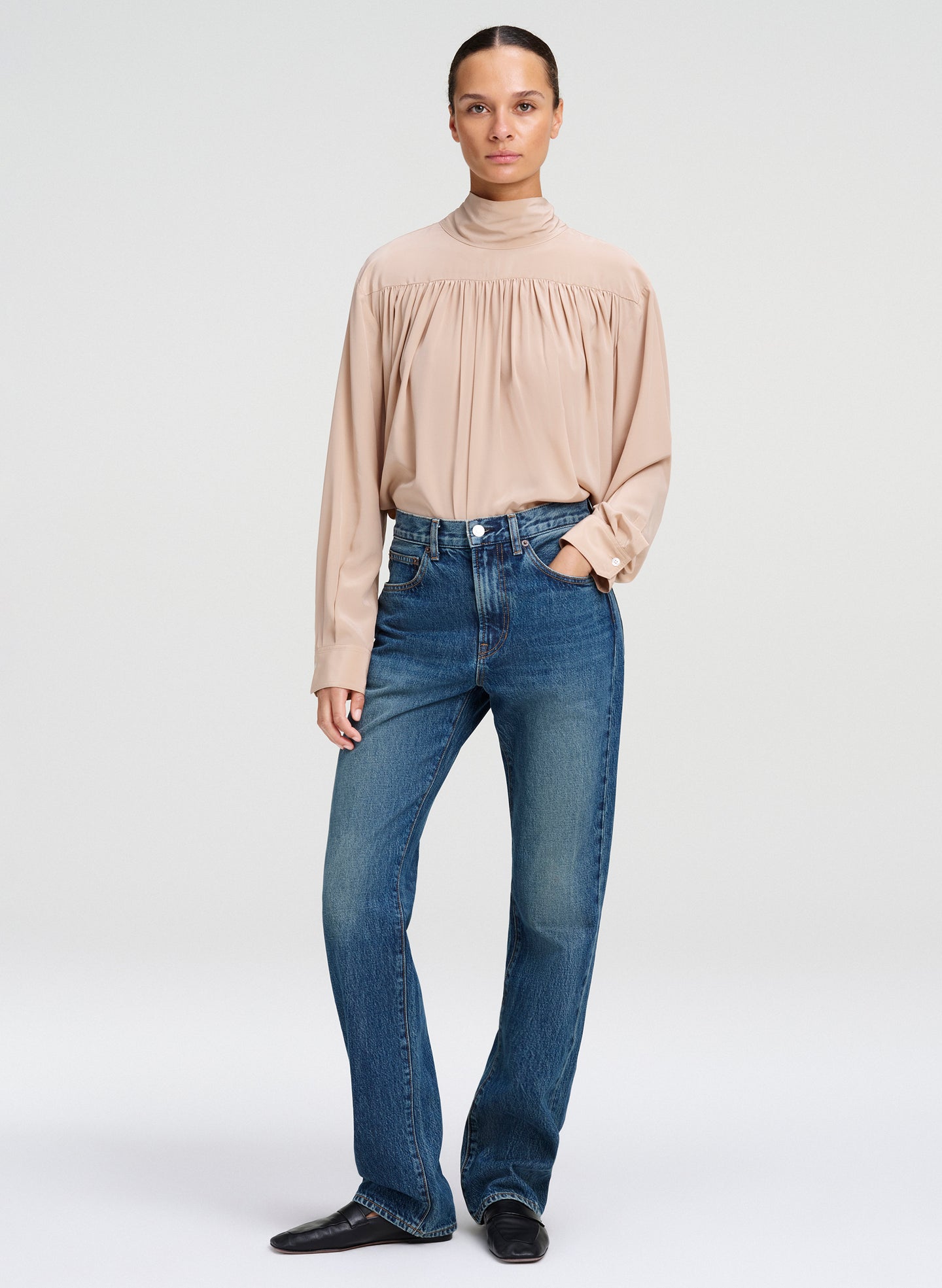 A model stands against a light gray studio background wearing a beige long-sleeve mock-neck blouse with gathered detailing at the chest, blue jeans, and black shoes.