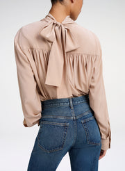 A woman shown from the back wearing a beige long-sleeve blouse with a large bow detail at the back neckline and blue jeans, against a plain light gray studio background.