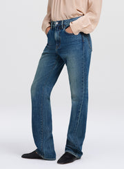 A person wearing medium-wash blue denim jeans with a straight-to-flare leg, paired with a light beige blouse and black shoes, shown from the waist down against a light gray studio background.