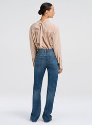 A woman shown from the back wearing a beige long-sleeve blouse with a tie-neck detail and blue jeans, photographed against a light gray studio background.