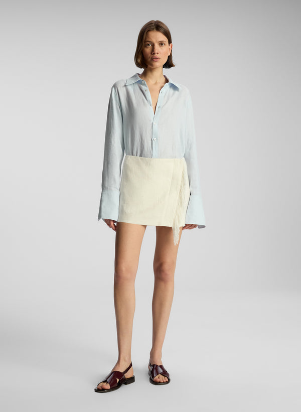 front view of woman wearing light blue button down and cream fringed mini skirt