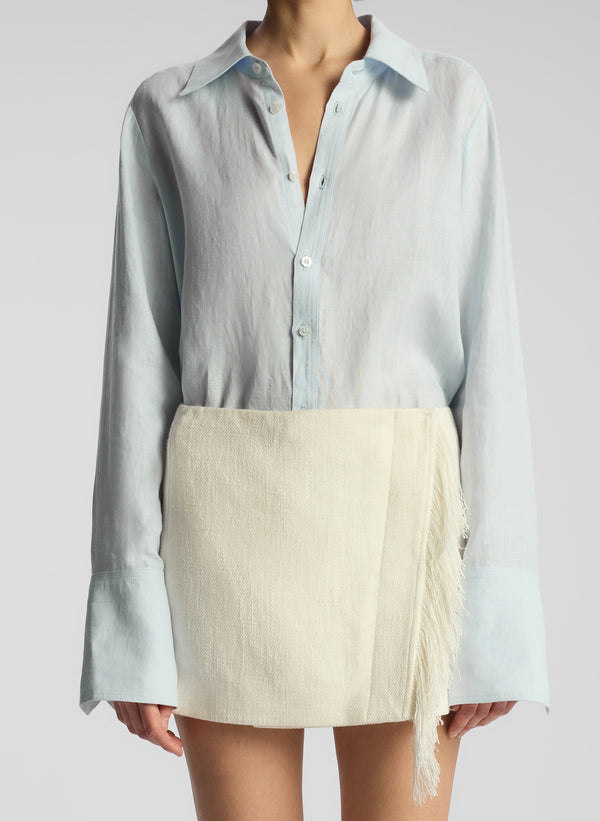 detail view of woman wearing light blue button down and cream fringed mini skirt