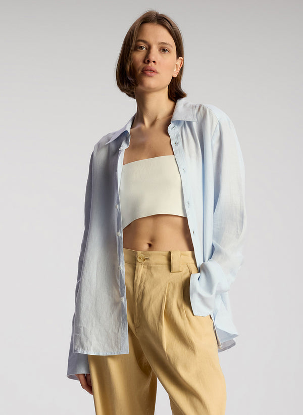 front view of woman wearing white cropped strapless top, light blue linen button down shirt and tan pants