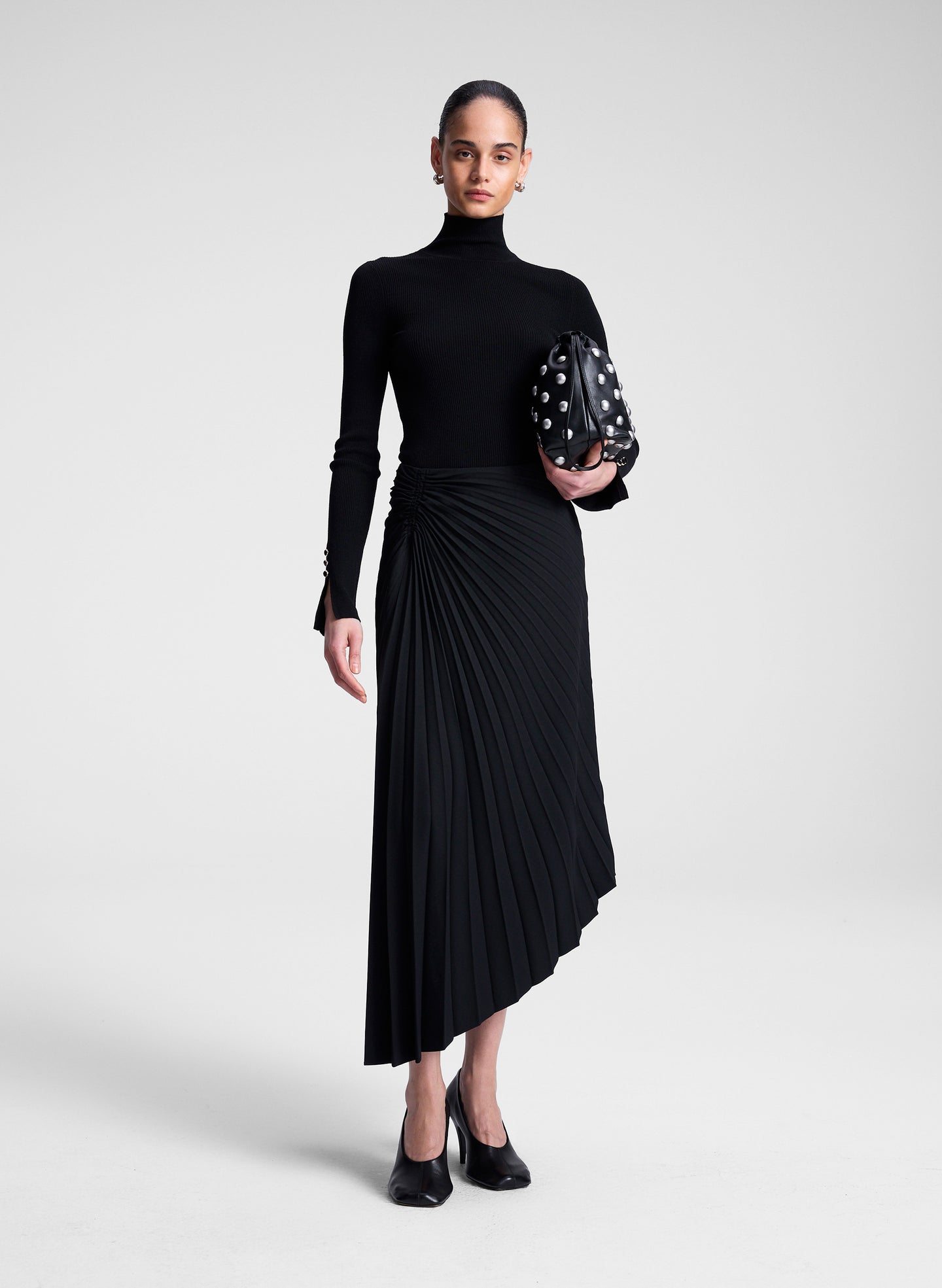 Woman wearing a black turtleneck and skirt holding a bag against a white background