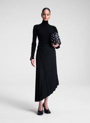 Woman wearing a black turtleneck and skirt holding a bag against a white background