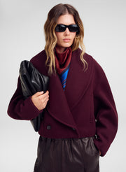 A model wearing a dark burgundy wool-blend wrap coat over a red  and blue striped scarf, holding a black leather clutch against a light gray studio background.