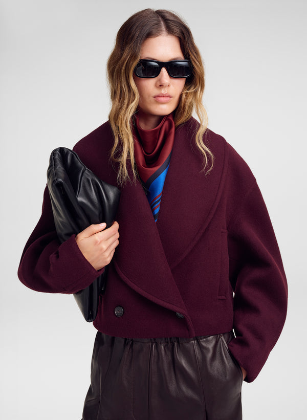 A model wearing a dark burgundy wool-blend wrap coat over a red  and blue striped scarf, holding a black leather clutch against a light gray studio background.