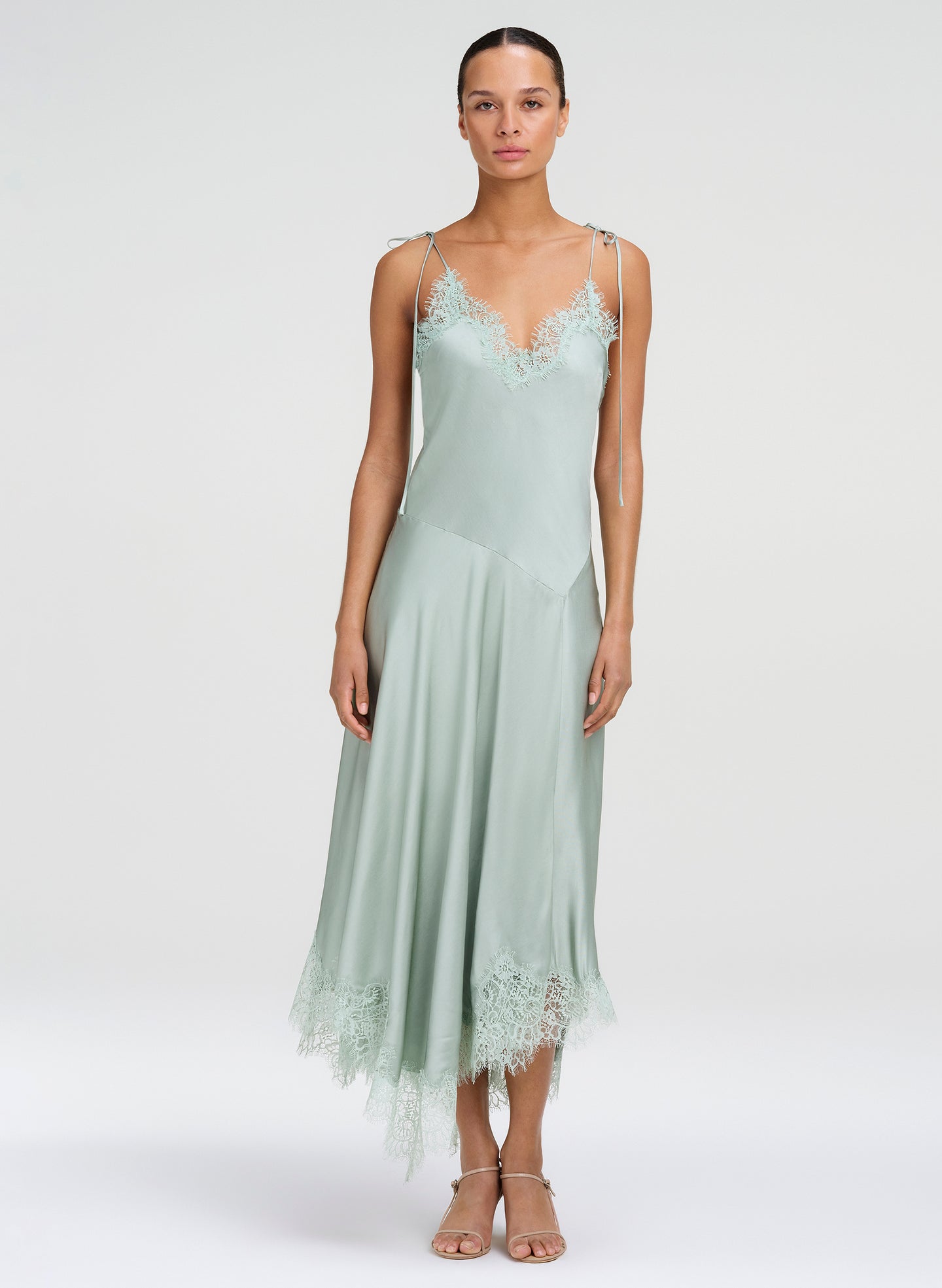 A model wearing a sage green satin slip dress with lace trim on the bodice and hem, paired with neutral sandals, against a light gray studio background.