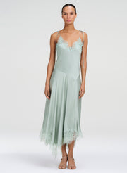 A model wearing a sage green satin slip dress with lace trim on the bodice and hem, paired with neutral sandals, against a light gray studio background.