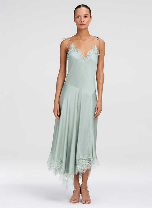 A model wearing a sage green satin slip dress with lace trim on the bodice and hem, paired with neutral sandals, against a light gray studio background.