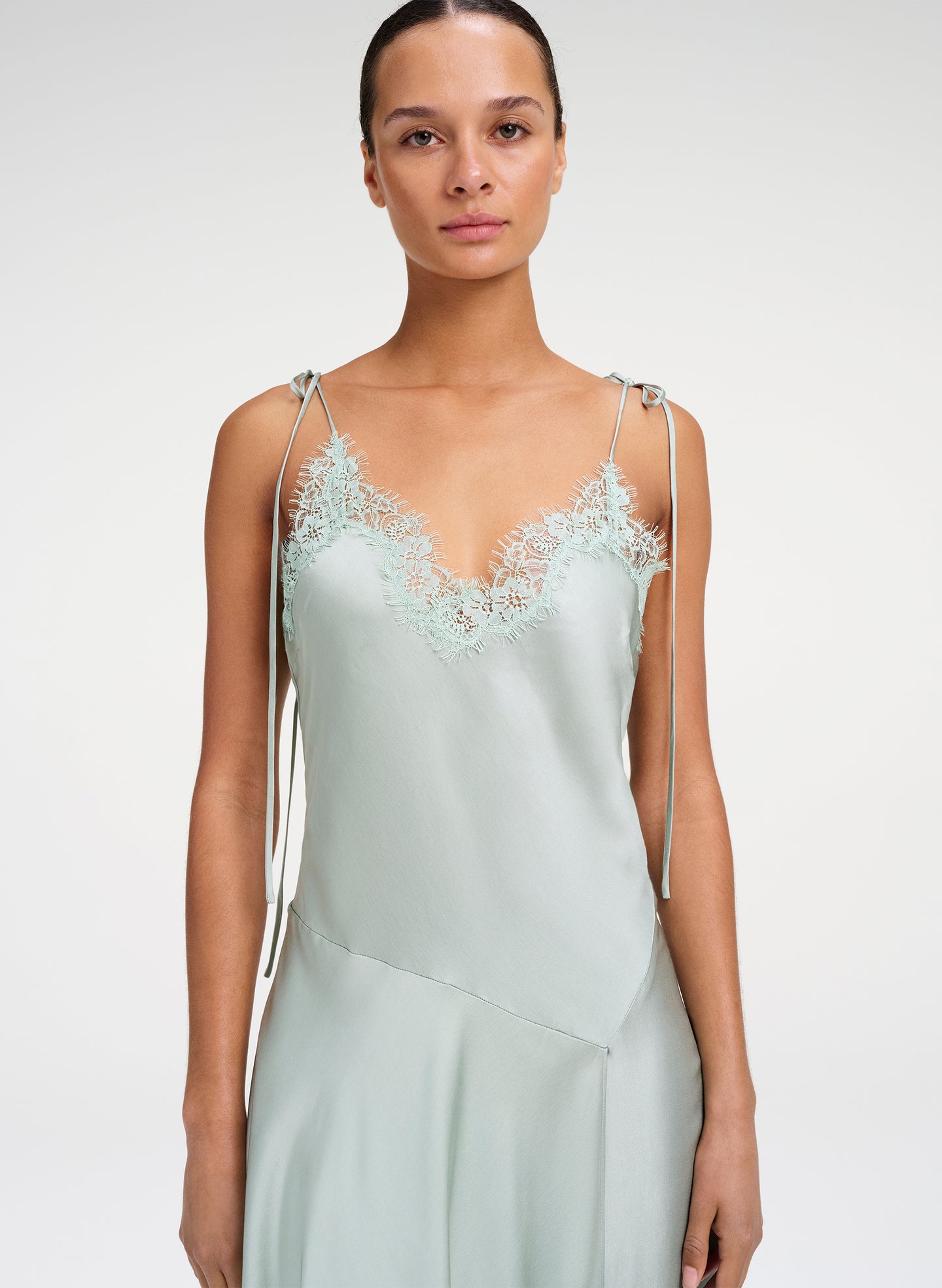 A model wearing a pale sage green satin slip dress with delicate lace detailing at the V-neckline and thin adjustable shoulder straps, set against a plain light gray studio background.