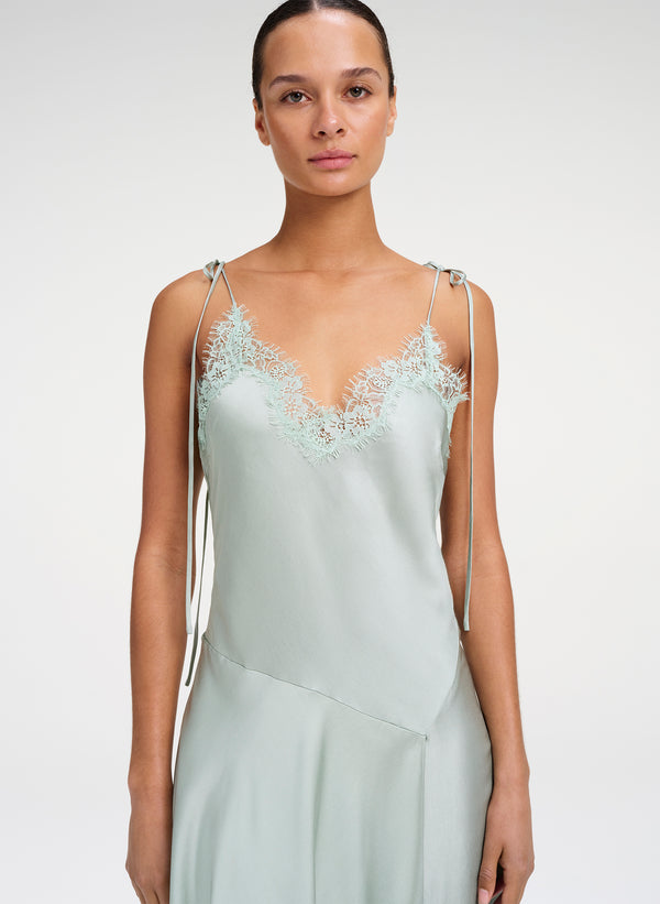 A model wearing a pale sage green satin slip dress with delicate lace detailing at the V-neckline and thin adjustable shoulder straps, set against a plain light gray studio background.