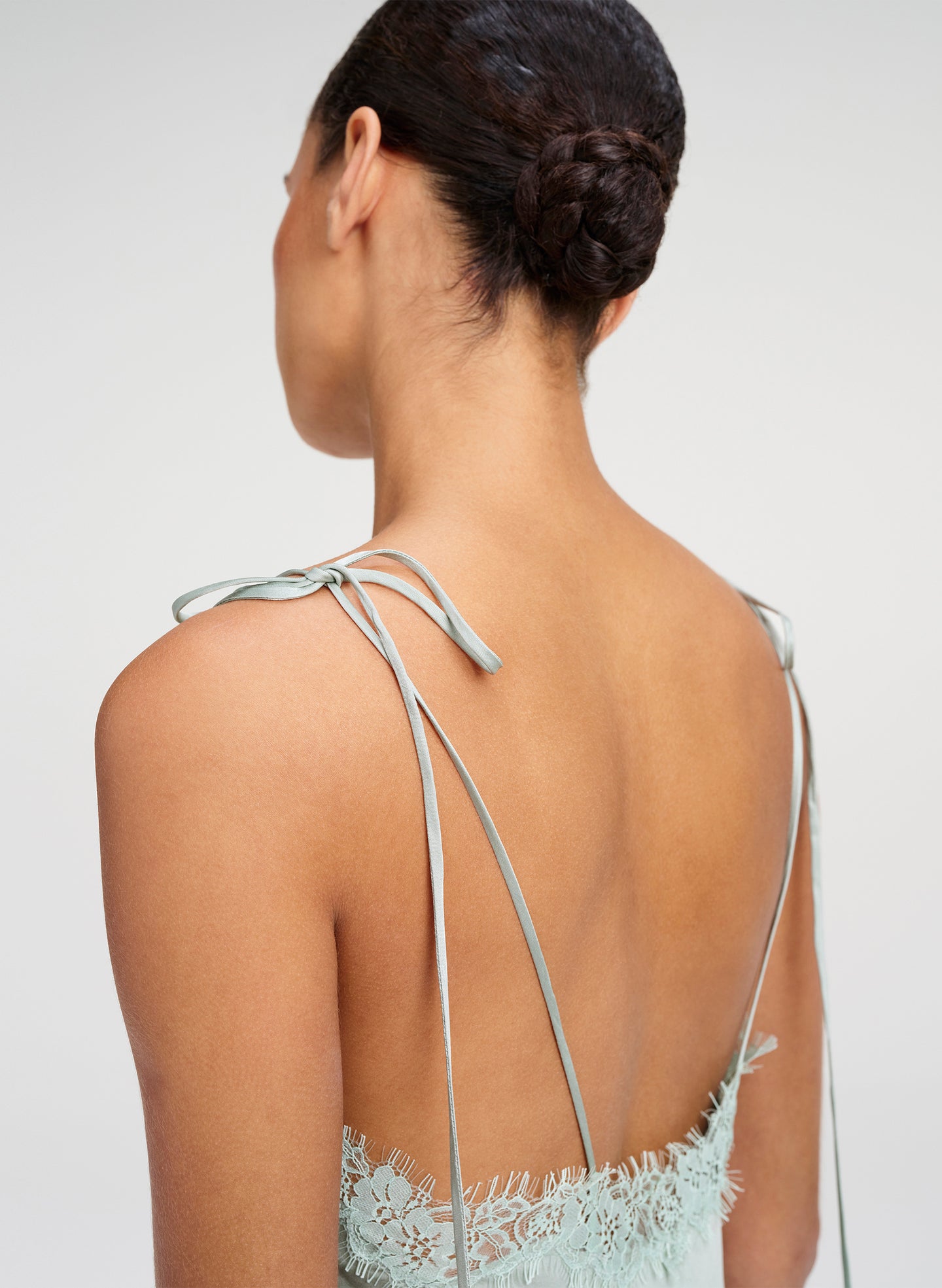 Rear view of a woman wearing a pale blue lace dress with thin tie-back straps, shown against a light gray studio background.